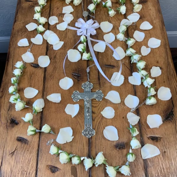 White rose petals and a rosary arranged around a silver cross on a wooden surface