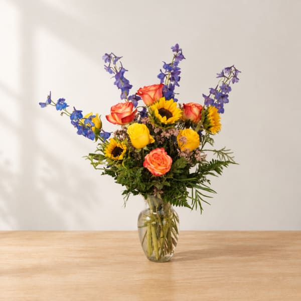 Colorful bouquet of roses, sunflowers, and blue delphinium in a glass vase