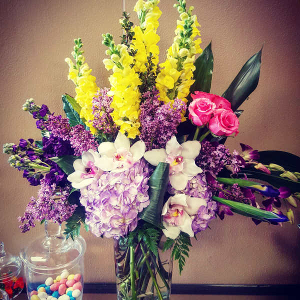 Mixed bouquet with yellow snapdragons, purple blooms, pink roses, and white orchids in a glass vase.