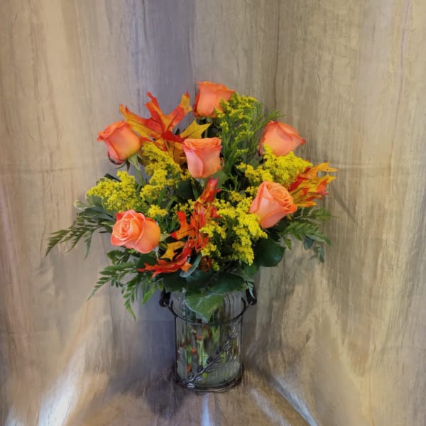 Orange roses in a glass vase with yellow filler flowers and autumn leaves