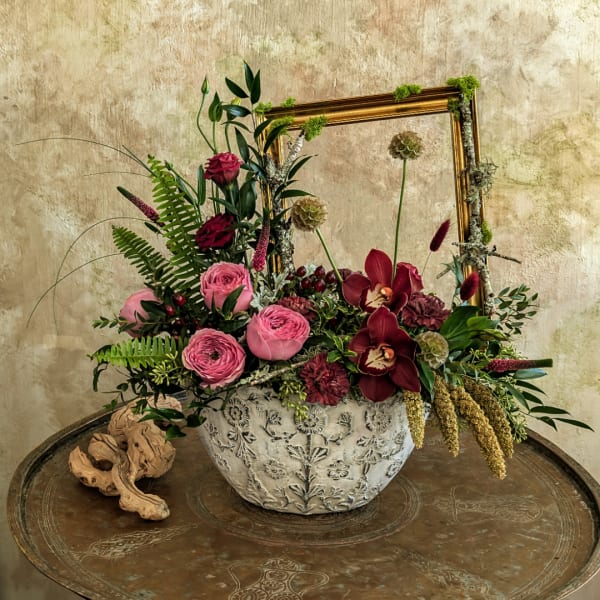 Pink and burgundy floral arrangement in a white ceramic bowl with a gold frame behind it