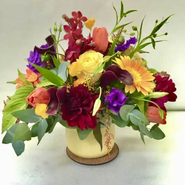 Colorful arrangement of yellow roses, gerbera daisies, tulips, and dark calla lilies in a cream pot.