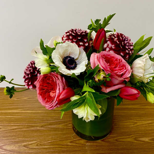Low pink and white flower arrangement with roses, anemones, tulips, and dahlias in a green glass cylinder vase