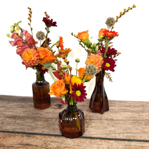 Three small floral arrangements in amber glass bottles on a wooden surface