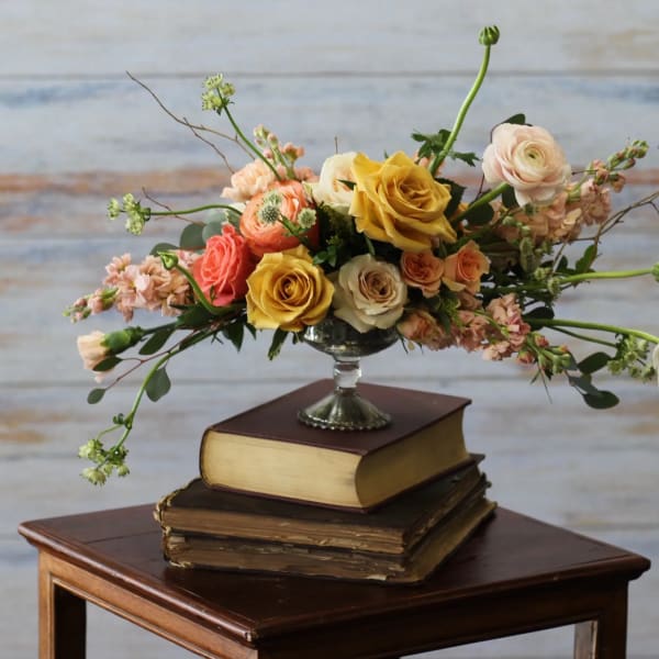 Low arrangement of yellow and peach roses and ranunculus in a silver pedestal bowl on stacked books.