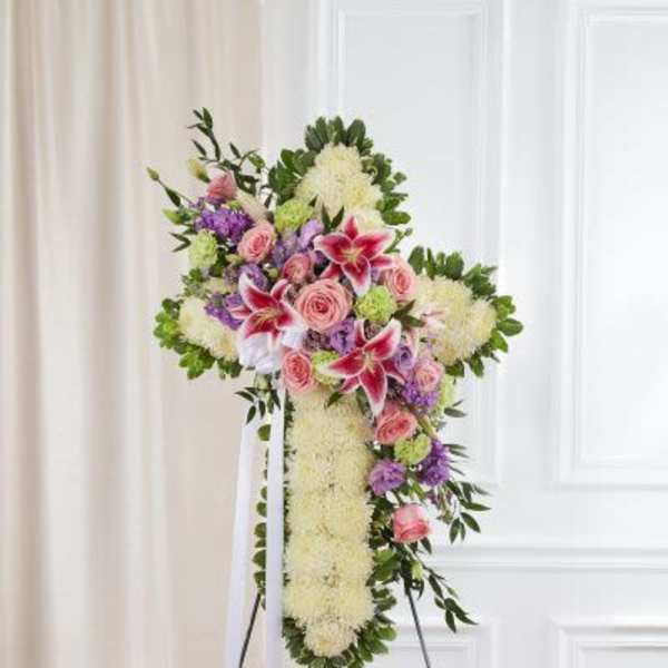 Standing floral cross with white base, pink roses, lilies, and lavender blooms on an easel