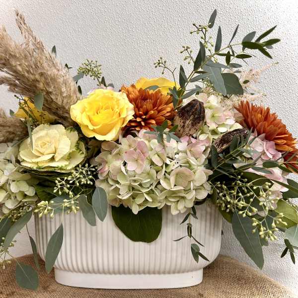 Mixed floral arrangement in a white ribbed container with yellow and orange blooms