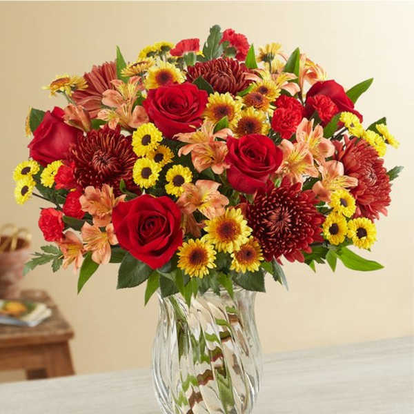 Mixed bouquet of red roses, yellow daisies, and orange flowers in a clear glass vase
