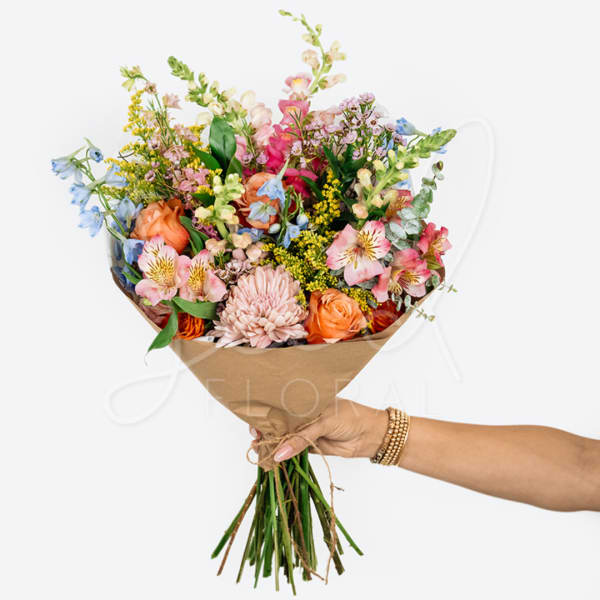 Handheld bouquet of mixed pink, peach, blue, and yellow flowers wrapped in brown paper
