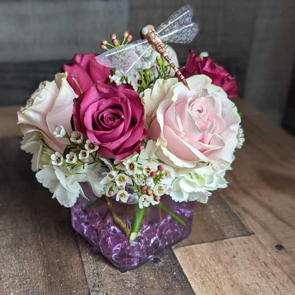 Short cube vase arrangement of pink and cream roses with white blooms and a decorative dragonfly pick.