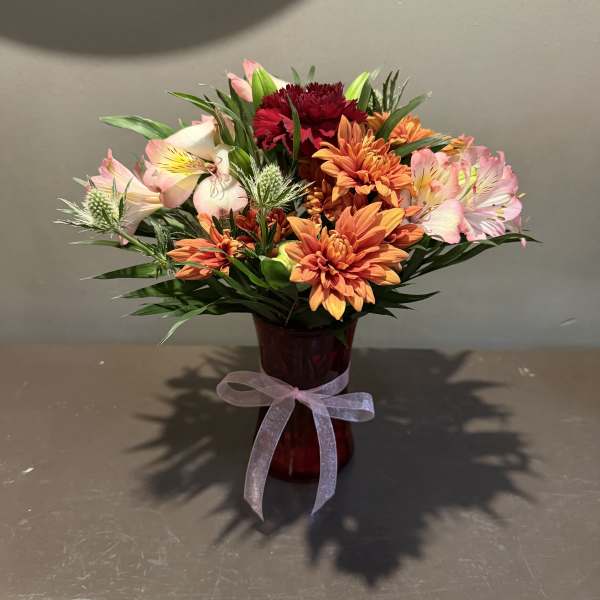 Mixed bouquet of orange, pink, and red flowers in a red vase with a ribbon