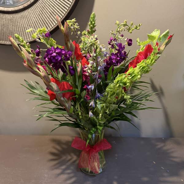 Tall mixed bouquet of red, purple, and green flowers in a glass vase with a pink ribbon.