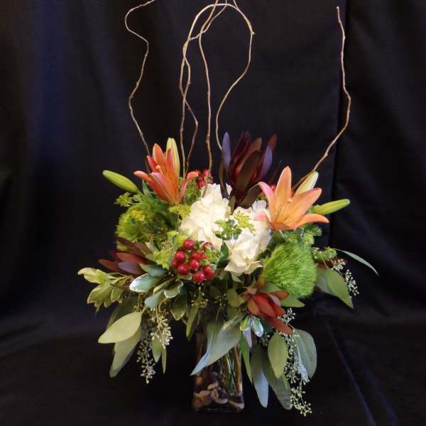 Tall floral arrangement with lilies, white hydrangea, and curly branches in a glass vase