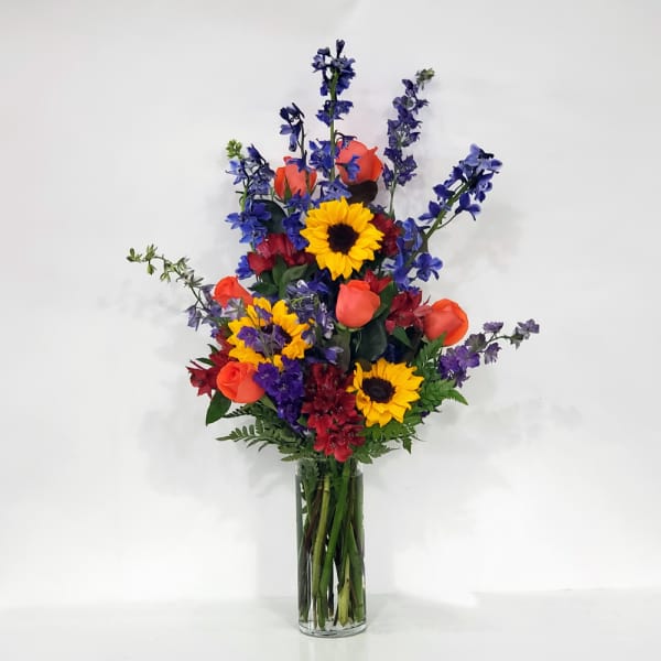 Tall bouquet of sunflowers, orange roses, and blue flowers in a glass vase