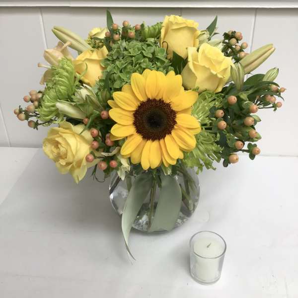 Yellow roses and a sunflower in a glass vase with a small candle nearby