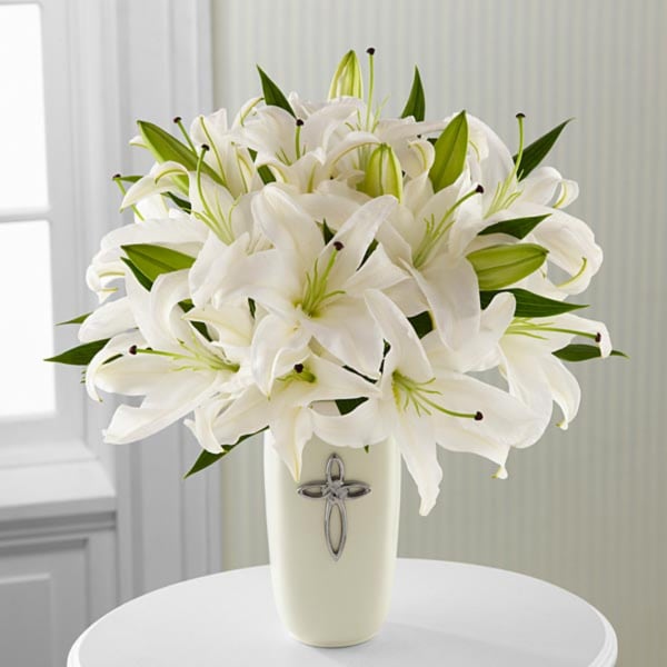 White lilies arranged in a cream vase with a silver cross emblem