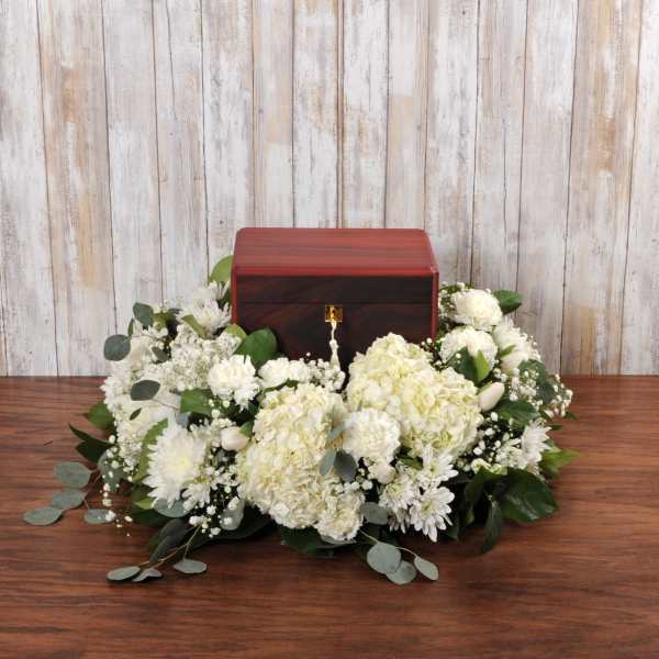 Wooden cremation urn surrounded by white hydrangeas, carnations, and mums on a table.