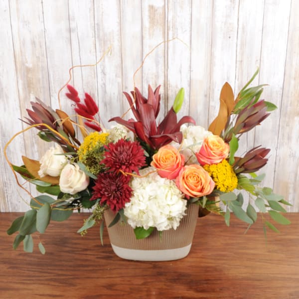 Low arrangement of orange roses, red lilies, burgundy mums, and white hydrangeas in a beige ceramic vase.