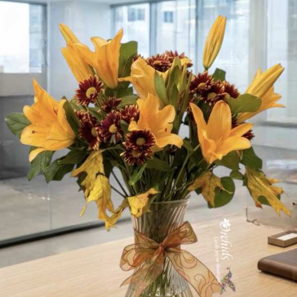 Orange lilies and burgundy daisies in a clear glass vase with a ribbon