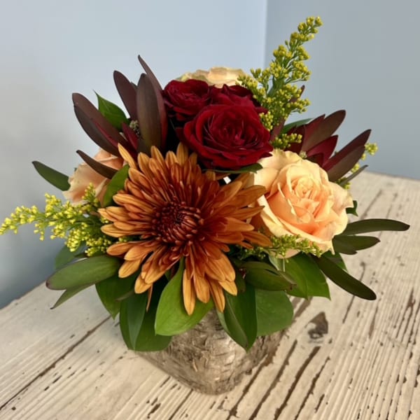 Compact bouquet of red and peach roses with an orange chrysanthemum in a birch container.