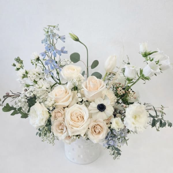 Low arrangement of ivory roses, white blooms, and pale blue flowers in a white ceramic vase