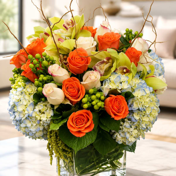 Orange roses and pale orchids with blue hydrangeas in a clear glass vase