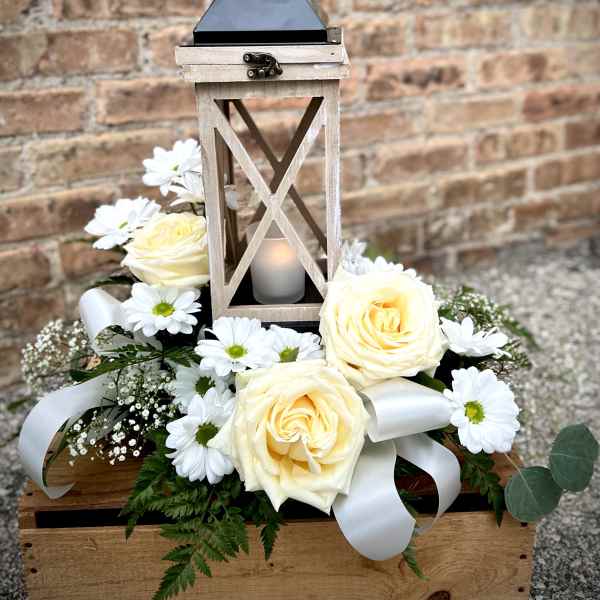 White roses and daisies arranged around a wooden lantern with a candle.