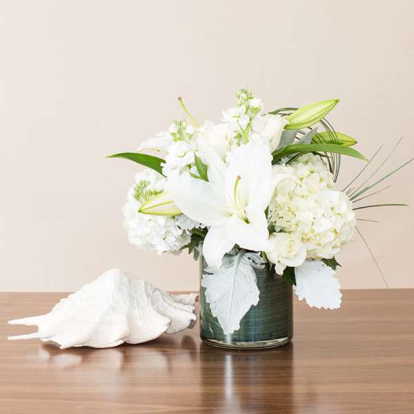 White lilies, roses, and hydrangeas in a glass vase beside a large white seashell.