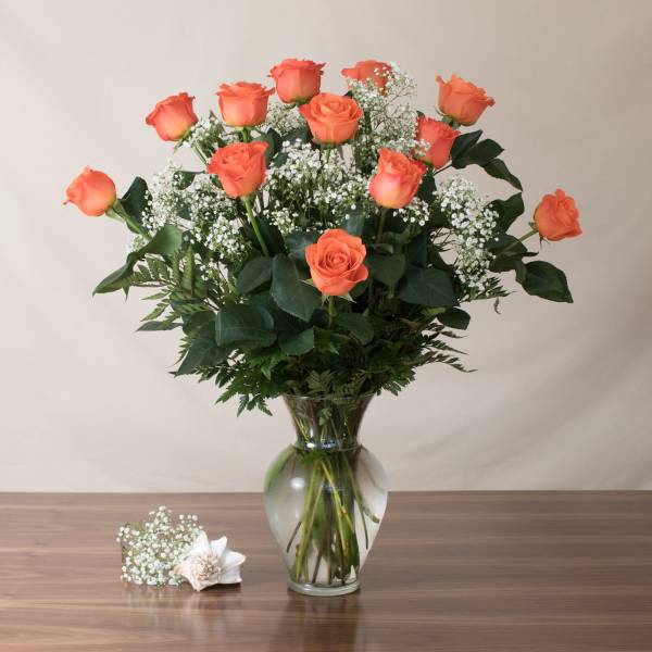Tall arrangement of orange roses and white filler flowers in a clear glass vase on a wooden table.