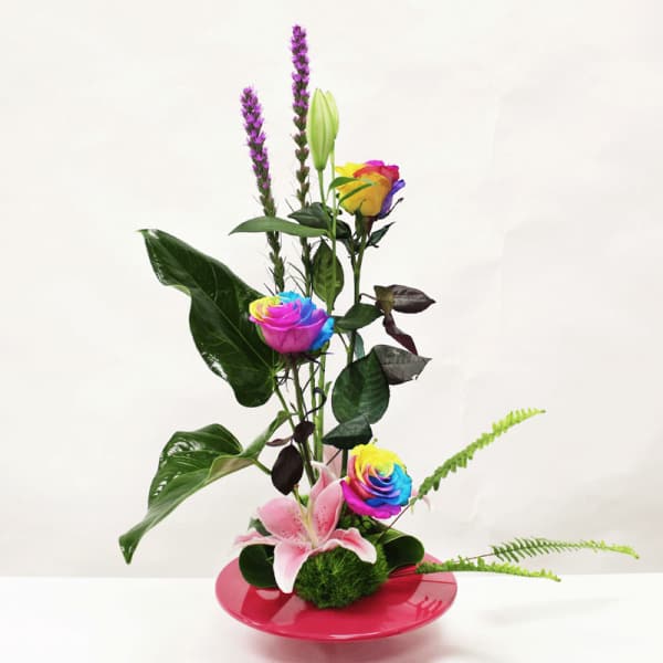 Modern arrangement with rainbow roses, pink lily, and purple spikes in a low pink dish