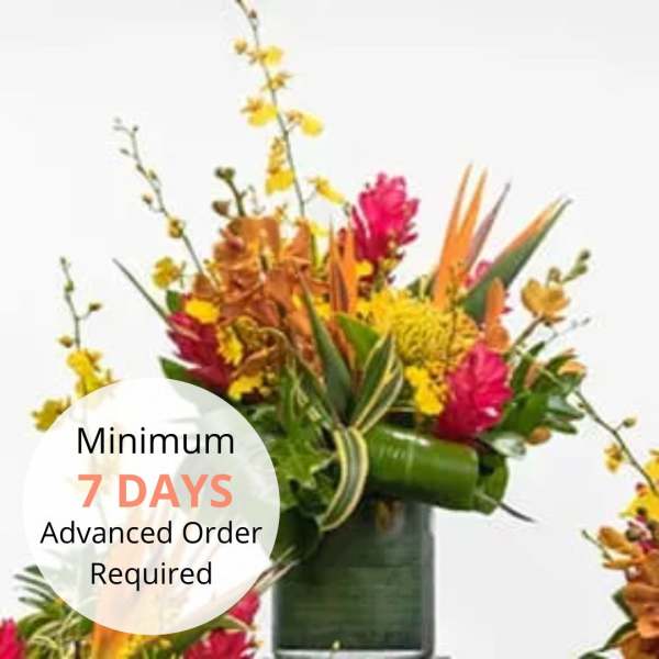 Tall tropical arrangement with yellow, orange, and pink flowers in a green cylinder vase