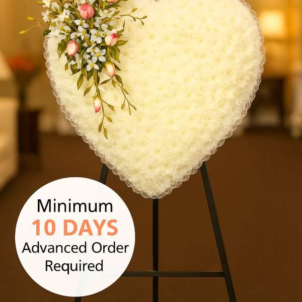 Heart-shaped white sympathy wreath with pink buds and small white flowers on a black easel stand indoors.