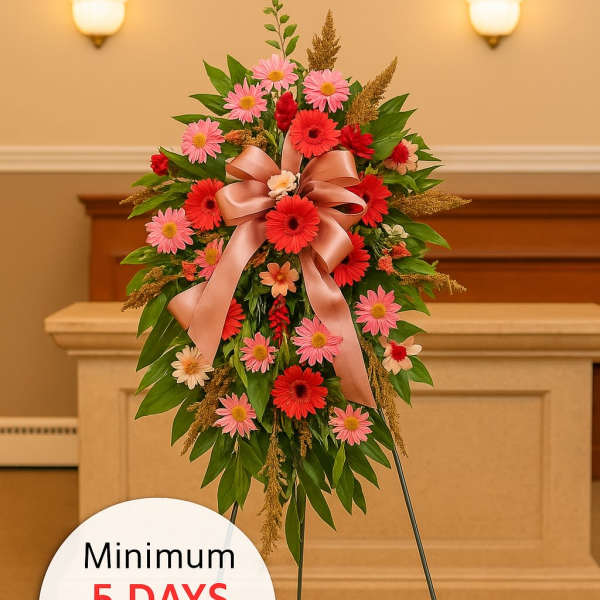 Tall sympathy spray of red and pink daisies with a peach bow on a metal easel in a chapel.