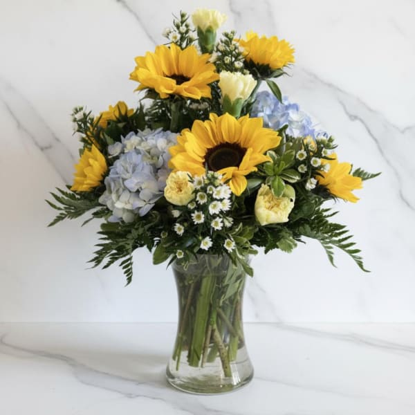 Bouquet of sunflowers, pale blue hydrangeas, and small white flowers in a clear glass vase