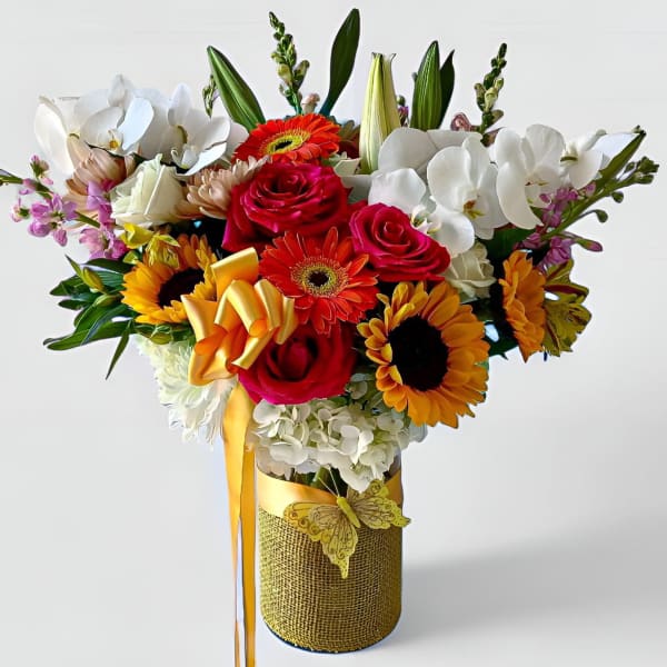 Tall mixed arrangement with sunflowers, red roses, orchids and gerberas in a yellow-wrapped vase with ribbon and butterfly