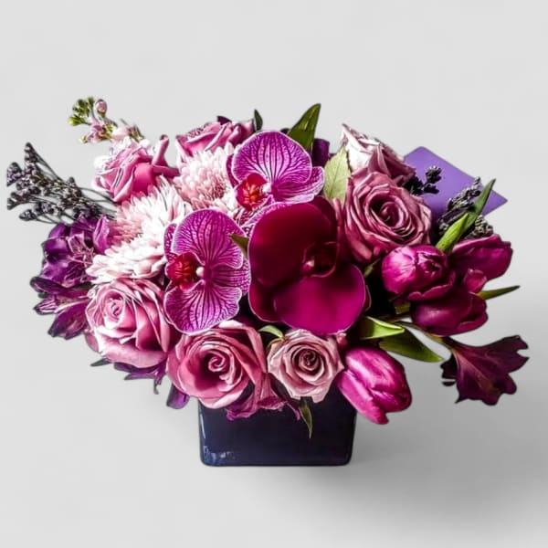 Low arrangement of purple orchids, pink roses, tulips, and mums in a dark square vase