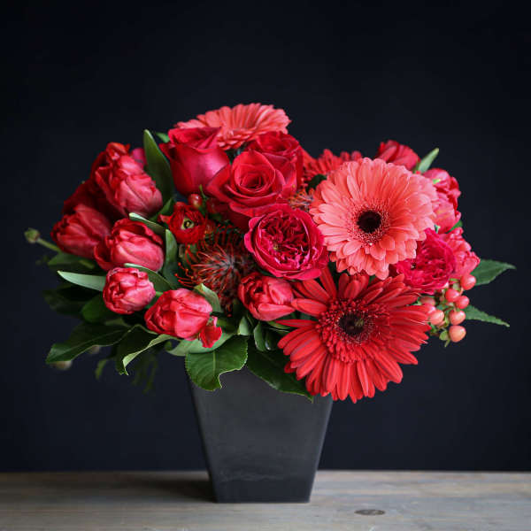Red and coral roses, gerbera daisies, and tulips arranged in a black vase.