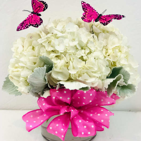 White hydrangea arrangement in a glass vase with pink ribbon and butterfly picks
