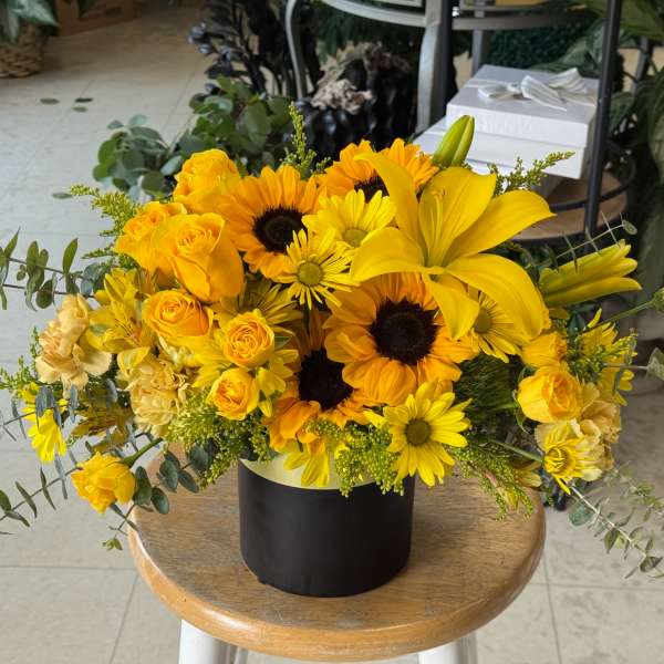 Bright yellow arrangement of sunflowers, lilies, roses, and daisies in a black cylinder vase