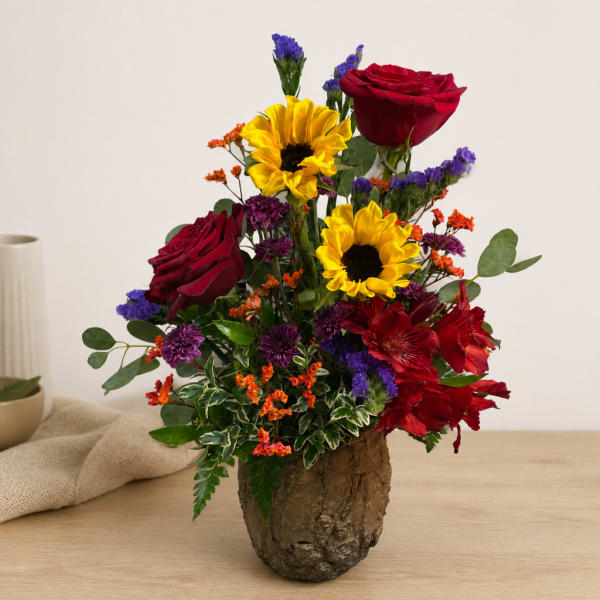 Mixed bouquet with red roses, yellow sunflowers, and purple accents in a rustic vase