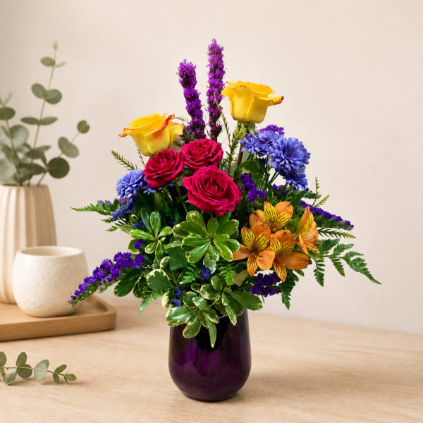 Colorful bouquet of roses, purple spikes, and blue flowers in a purple vase