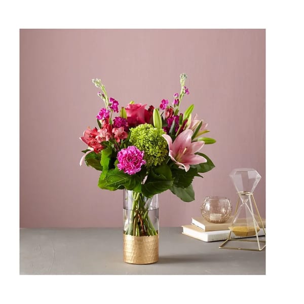 Mixed pink lilies, roses, carnations, and green hydrangea in a clear vase with gold base on a tabletop