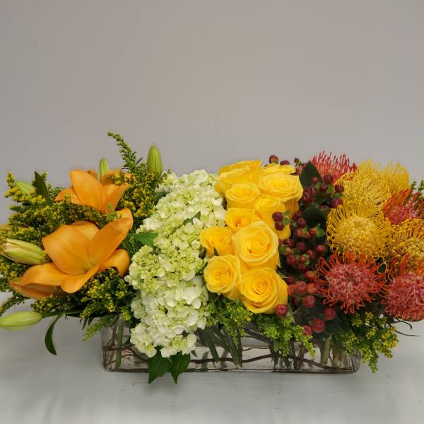 Long floral centerpiece with orange lilies, yellow roses, and mixed blooms in a rectangular vase