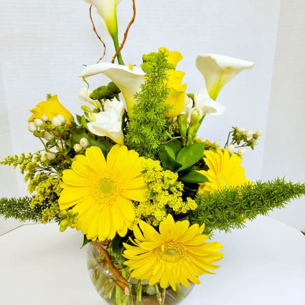Compact arrangement of yellow gerbera daisies and white calla lilies in a clear round vase