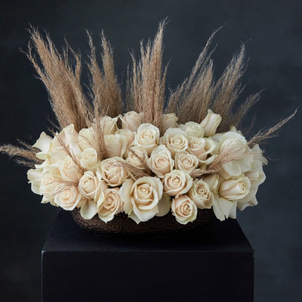 Cream rose arrangement with tall dried grass accents in a dark bowl