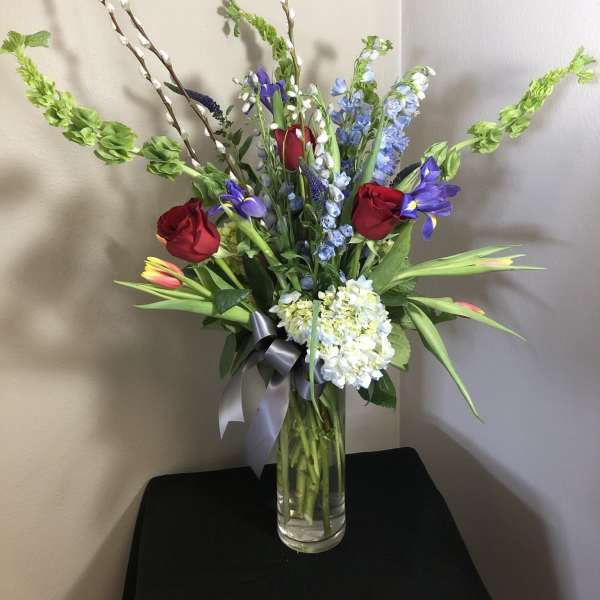Tall mixed bouquet in a clear glass vase with red roses, blue flowers, and pussy willow stems