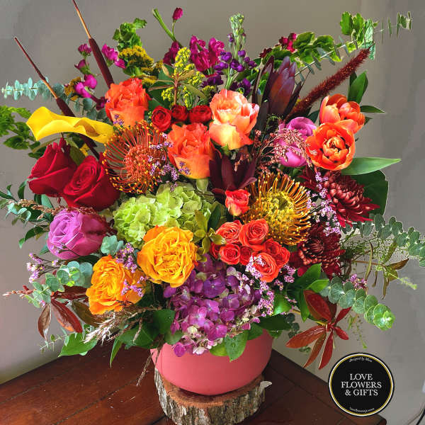 Large colorful arrangement of roses, hydrangeas, protea and mixed blooms in a pink vase on a wood slice