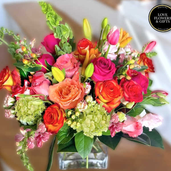 Vibrant arrangement of pink, orange, and yellow roses with lilies and green hydrangeas in a clear glass vase.