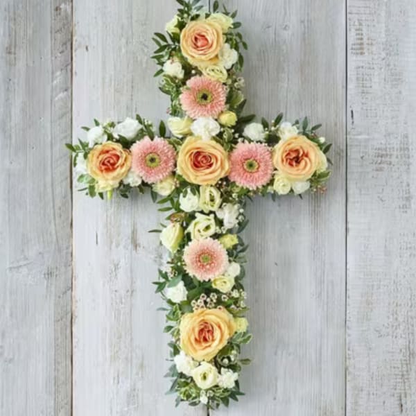 Floral cross arrangement with peach roses and pink daisies on a white wood background