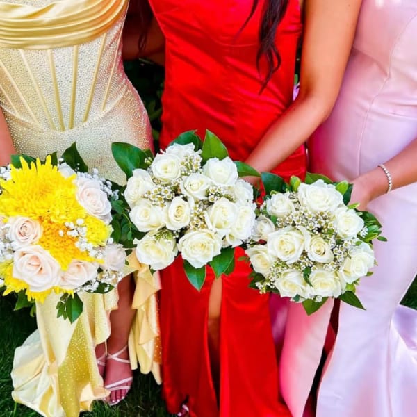 Three women hold bouquets of white and yellow roses in formal dresses.